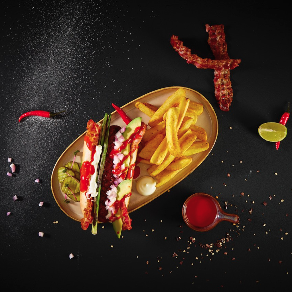 Hot-Dog-Bun mit gegrillter Wurst, bestrichen mit veganer bedda Mayo, belegt mit Avocado-Scheiben, gerösteter Frühlingszwiebel, roten Zwiebeln, Gewürzgurke, krossem Bacon und Chiliringen, großzügig mit Fire Cracker Sauce übergossen